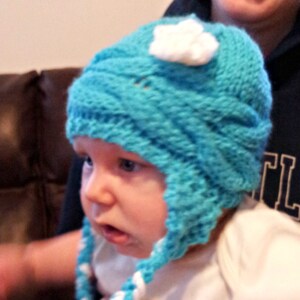 May include: A baby wearing a blue knitted hat with a white flower on top. The hat has white braided ties that are tied under the chin. The baby is wearing a white shirt.