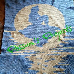 May include: A blue crocheted blanket with a white crocheted silhouette of a mermaid sitting on a rock under a full moon. The text "Possum's Projects" is written in green across the image.