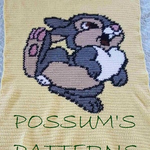May include: A yellow crochet blanket featuring a grey and brown bunny rabbit design. The blanket is labelled "POSSUM'S PATTERNS" in green lettering.