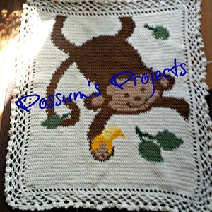 Könnte beinhalten: Eine gehäkelte Babydecke mit einem braunen Affenmotiv, grünen Blättern und einer gelben Banane. Die Decke hat einen weißen Häkelrand.