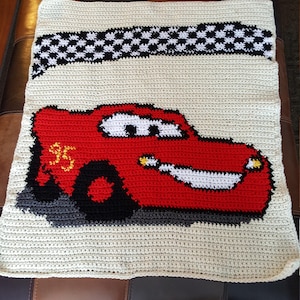 Könnte beinhalten: Häkeldecke mit einem roten Comic-Rennwagen mit der Nummer 95 an der Seite. Das Auto befindet sich auf einem weißen Hintergrund mit einem schwarz-weißen Karostreifen oben.