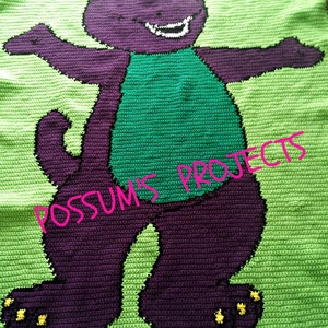 May include: A handmade crochet blanket featuring a purple dinosaur character with a green belly and a smiling face. The blanket is primarily green with the dinosaur in shades of purple and green. The text "POSSUM'S PROJECTS" is written across the blanket.