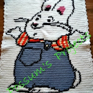 Puede incluir: Una manta de crochet blanca con un conejo de dibujos animados que lleva un mono y una camisa a rayas. El conejo tiene orejas rosas y una nariz negra. La manta tiene un borde festoneado.