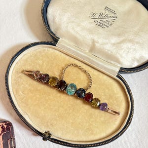 Può includere: Una spilla a barra color oro con una fila di nove pietre preziose ovali sfaccettate multicolori nelle tonalità del viola, giallo, rosso scuro e blu, dotata di una sottile catenella di sicurezza, riposta in un portagioie ovale foderato in velluto blu navy.