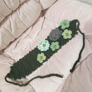May include: Dark green crochet headband adorned with several crochet flowers in shades of green and gray. The flowers are accented with wooden buttons. The headband is lying on a pink, textured surface.