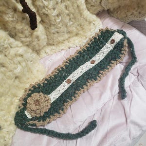 May include: Handmade crochet headband in green, tan, and white. The headband features a floral embellishment, a white lace stripe, and three brown buttons. The edges are scalloped, and the ties are green.