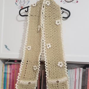 May include: A beige crochet scarf with white lace trim and flower accents. The scarf is hanging on a black hanger. The scarf has a textured pattern and is decorated with small, white, crocheted flowers and a lace trim.