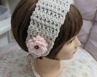 May include: A light gray crocheted headband featuring a textured stitch pattern and a soft pink crocheted flower accent on the side. The flower has a small dark brown button at its center. The headband includes thin crocheted ties at the base.