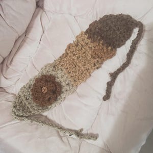 May include: A handmade crochet headband in shades of gray, tan, and brown. The headband features a flower with a wooden button center. The design includes a long, braided tie for adjusting the fit. The item is displayed on a white surface.
