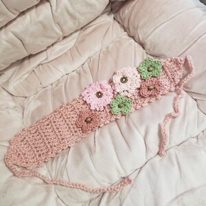 May include: A handmade, light pink crochet headband adorned with five crocheted flowers in shades of pink, green, and brown. The flowers have brown button centers. The headband has a braided tie on each end.