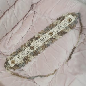 May include: A handmade crochet headband in neutral tones of beige, gray, and brown. The headband features a cream-colored lace panel down the center, adorned with four wooden buttons. The ends of the headband have long, crocheted ties.