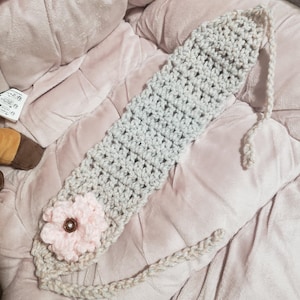 May include: A handmade crochet headband in light gray, featuring a light pink flower with a brown center. The headband is lying on a pink, plush surface. The headband is designed to be worn around the head.