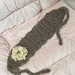 May include: A handmade, dark gray crochet headband with a cream-colored flower accent. The headband is made of thick yarn and features a button in the center of the flower. The ends of the headband are designed to tie.