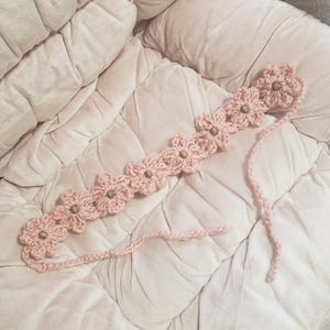 May include: A pink crochet headband featuring a series of flower designs with wooden bead centers. The headband is made of a soft, textured yarn and is shown on a pink quilted surface.