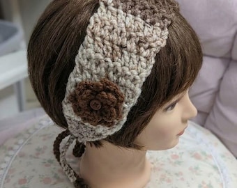 May include: A crochet headband featuring a color-blocked design in shades of brown and cream. The accessory includes a dark brown crocheted flower embellishment with a central button and two braided ties hanging from the bottom edge.