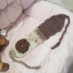 May include: A handmade crochet headband in shades of brown and cream. The headband features a large, dark brown flower with a button center. The design includes a long, braided tie on one end.