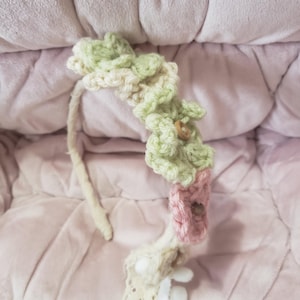 May include: A handmade headband with a crochet floral design. The flowers are in shades of green and pink, with a small wooden button detail. The headband also features a lace and pom-pom trim.