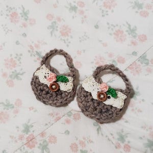 May include: Two small, handmade crochet baskets in a muted brown color. Each basket features a white lace trim, a pink flower, a green leaf, and a wooden button. The baskets have small handles and are set against a floral patterned background.