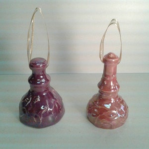 May include: Two ceramic bottle stoppers with iridescent glaze. The stoppers have a round base and a tall, slender neck with a loop handle. One stopper is a deep purple color and the other is a light brown color.