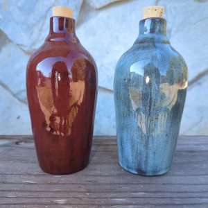 May include: Two ceramic bottles with cork stoppers. One is a deep reddish-brown, the other is blue with brown streaks. Both have a glossy finish and a rounded shape. The bottles are displayed on a wooden surface.