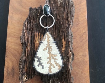 Puede incluir: Un collar de plata con un colgante en forma de lágrima que presenta una piedra de ágata dendrítica blanca y dorada. El colgante está unido a una cadena de plata con una piedra ovalada negra.