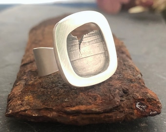 May include: A silver ring with a square-shaped design featuring a cutout in the center. The cutout reveals a textured, silver surface with a crack running through it.