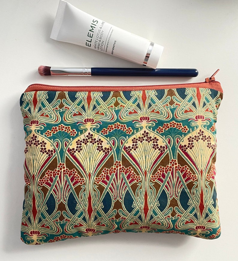 May include: A cosmetic bag with a colorful floral pattern in teal, brown, and red, with a rust-colored zipper. A white tube of Elemis lotion and a makeup brush are on top of the bag.