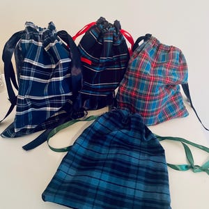 May include: Five drawstring bags in different tartan patterns. The bags display blue, red, and black plaid designs. Each bag has a contrasting ribbon drawstring closure. The bags are arranged on a white surface.