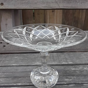 May include: A clear glass pedestal cake stand with a cut glass design. The stand has a round base and a tall, slender stem.