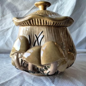May include: A ceramic mushroom-shaped cookie jar with a lid. The jar is tan with three-dimensional mushroom details and a textured surface. The lid has a mushroom-shaped knob. The jar has cut-out grass details.