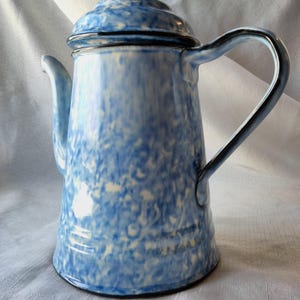 May include: A blue and white speckled enamelware coffee pot with a lid and handle. The pot has a classic design with a spout for pouring. The handle and rim are a darker shade of blue. The pot is likely vintage.