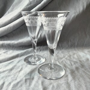 May include: Two clear glass shot glasses with a white etched design around the top rim. The glasses are on a white fabric background.