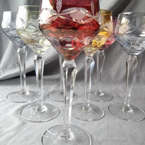 May include: A collection of six crystal wine glasses with colored bowls. The glasses have clear stems and bases. The bowls are various colors, including red, yellow, and clear, with etched designs. The glasses are arranged on a light gray surface.
