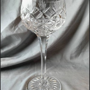 May include: A clear crystal wine glass with a long stem and a round base. The bowl of the glass features a diamond-cut pattern, and the base has a starburst design. The glass is designed for serving wine.