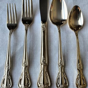 May include: A set of gold-colored flatware, including forks, a knife, and spoons, arranged on a white surface. The utensils have ornate floral detailing on the handles. The knife blade is a darker silver color.