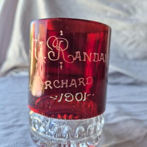 May include: A ruby red glass with etched white lettering. The glass has a clear, patterned base and a cylindrical top. The text reads "R. ANDA", "ORCHARD", and "1901". The glass is likely a vintage collectible.