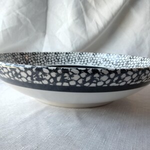 May include: White ceramic bowl with a black and white spotted rim and a solid black band. The bowl has a wide, shallow design and a white interior. This decorative bowl is suitable for serving.