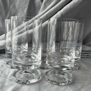 May include: Set of five clear glass tumblers with a short stem and a wide base. The glasses are arranged in a row on a white fabric surface.
