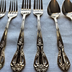 May include: Close-up of a set of gold-colored silverware, including forks and spoons. The silverware has ornate floral detailing on the handles. The utensils are arranged on a light gray, textured surface.