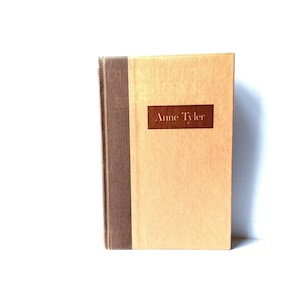 May include: A hardcover book with a light brown cover and a dark brown spine. The title "Anne Tyler" is printed in dark brown on a rectangular label on the front cover. The book is set against a white background.
