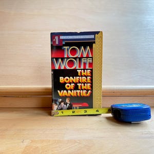 May include: A paperback book titled The Bonfire of the Vanities by Tom Wolfe with a black cover featuring yellow and red text, a skyscraper graphic, and a small illustration of two people. A yellow measuring tape shows a width of 4 inches.