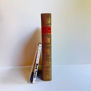 May include: A tall, olive-green hardcover book titled The Innocents Abroad by Mark Twain with gold-embossed illustrations on the spine, positioned next to a thin, white paperback book titled The Wit & Wisdom of Mark Twain with black text.