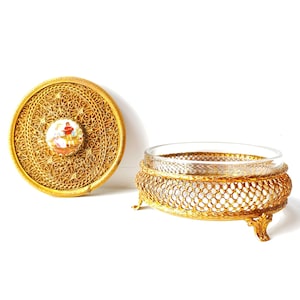 May include: A gold-colored metal trinket box with a glass insert and a decorative lid. The lid features a delicate filigree design and a small porcelain cameo with a painted scene.