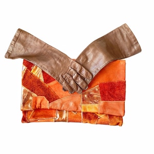 May include: A pair of brown leather gloves folded over a patchwork clutch purse. The clutch is made of different shades of orange and brown suede.