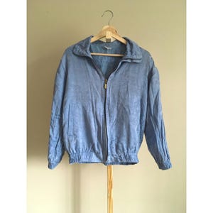 May include: A light blue jacket with a floral pattern, a zip-up front, and a stand-up collar. The jacket has a ribbed hem and cuffs. The word "FUDA" is visible on the inside of the collar. The jacket is hanging on a wooden hanger.