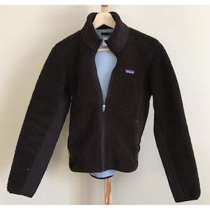 May include: A dark brown fleece jacket with a full-length zipper and a high collar. The jacket has a small purple and white logo patch on the chest. The sleeves have a darker brown panel down the sides. The jacket is hanging on a wooden hanger.