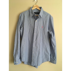 May include: A long-sleeved button-down shirt in a blue and white checkered pattern. The shirt has a classic collar and cuffs, with a small pink logo on the chest. The shirt is hanging on a wooden hanger.