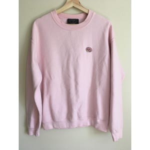 May include: A light pink sweatshirt with a crew neck and long sleeves. A small, circular logo is embroidered on the left chest. The brand name is printed on a tag at the back of the neck. The sweatshirt is hanging on a wooden hanger.