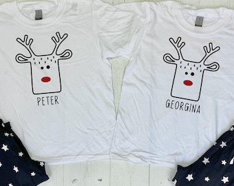 May include: Two white t-shirts with a reindeer design and the names "PETER" and "GEORGINA". They are paired with navy blue pajama bottoms with a white star pattern. A matching festive set.