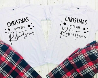 May include: Two sets of Christmas pyjamas. Each set features a white t-shirt printed with "Christmas with the Robertsons" and black stars. The pyjama bottoms are red, black, and white tartan plaid.